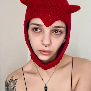 Könnte beinhalten: Eine Person trägt eine rote, gehäkelte Teufelskappe mit Hörnern. Die Mütze hat eine spitze Spitze und bedeckt Kopf und Hals der Person. Die Person trägt auch ein schwarzes Tanktop und eine silberne Kettenkette mit einem violetten Kristallanhänger.