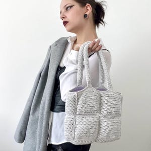 Può includere: Una borsa tote in crochet grigia con un designo quadrato. La borsa ha due manici e una fodera di colore lilla chiaro.