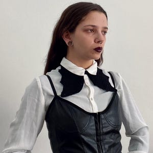 May include: A person wearing a white button-down shirt with a black crocheted collar, a black leather corset, and a black belt with silver eyelets.