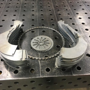 Op de afbeelding: Een close-up van een metalen tafel met een cirkelvormig metalen stuk en verschillende gestapelde metalen stukken in de vorm van een halve maan. De tafel heeft een rasterpatroon van gaten.
