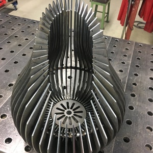 Könnte beinhalten: Eine Metallsculptur aus mehreren dünnen Metallteilen, die zu einer Birnenform zusammengeschweißt sind. Die Skulptur hat eine kreisförmige Öffnung in der Mitte und Zahlen auf der Außenseite der Metallteile.