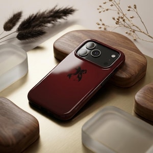 Può includere: Una cover per smartphone color bordeaux intenso con un piccolo fiocco nero centrato. La cover presenta una finitura lucida e una sezione rialzata per la fotocamera che ospita tre lenti circolari e un flash, mantenendo un profilo pulito.