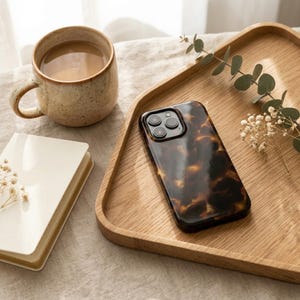 Può includere: Una custodia per smartphone con un motivo tartarugato lucido marrone e nero con un ritaglio per la tripla fotocamera, appoggiata su un vassoio di legno chiaro accanto a una tazza in ceramica piena di caffè e un piccolo taccuino bianco con fiori secchi.