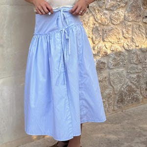 Puede incluir: Una falda midi a rayas azules y blancas con cintura anudada. La falda se combina con un top color crema y sandalias marrones. La falda tiene una silueta amplia y fluida.