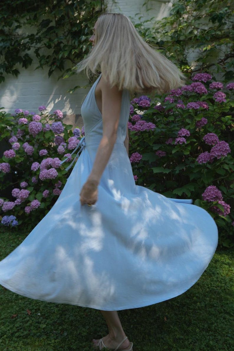 May include: A light blue sleeveless wrap dress with a V-neckline and a tie waist. The dress features a full, flared skirt that is captured in motion, creating a wide, circular silhouette that extends outward from the waistline.