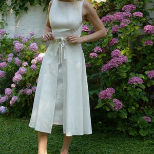 May include: A sleeveless off-white midi dress featuring a round neckline, a fitted bodice, and a waist tie that secures at the front. The skirt has a split front design, and the garment is paired with light-colored strappy block-heeled sandals.