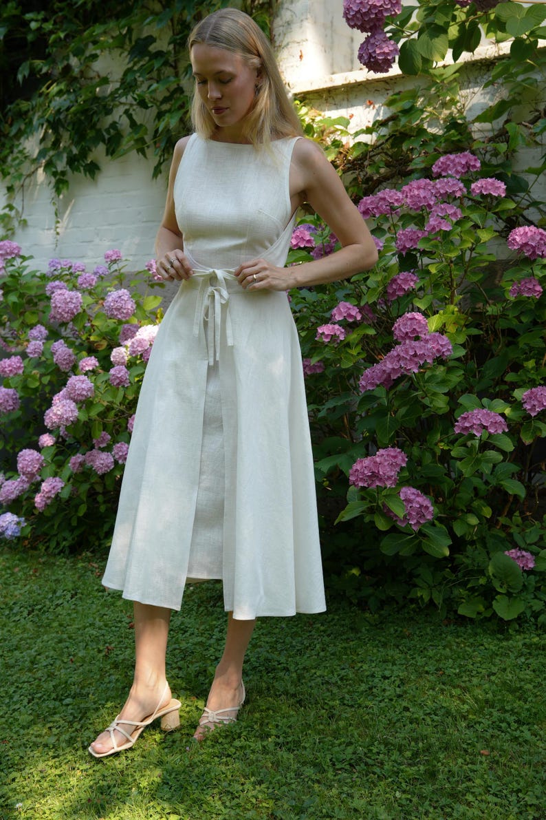 May include: A sleeveless off-white midi dress featuring a round neckline, a fitted bodice, and a waist tie that secures at the front. The skirt has a split front design, and the garment is paired with light-colored strappy block-heeled sandals.