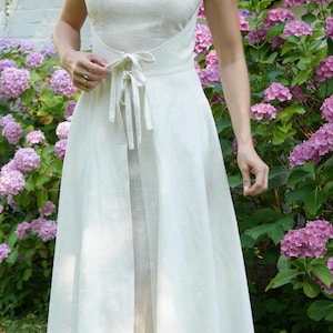 May include: A sleeveless off-white midi dress featuring a high neckline and a fitted bodice. The waist is accented with a matching fabric tie that secures in the front, leading into a flared skirt with a central vertical opening at the hemline.