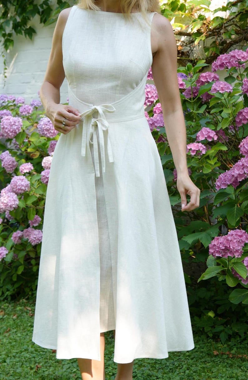 May include: A sleeveless off-white midi dress featuring a high neckline and a fitted bodice. The waist is accented with a matching fabric tie that secures in the front, leading into a flared skirt with a central vertical opening at the hemline.