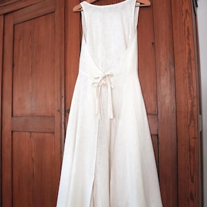 May include: A sleeveless off-white dress featuring a deep open back design with a tie closure at the waist. The dress has a flared skirt that falls below the knee and is constructed from a textured fabric, hanging from a wooden clothes hanger.