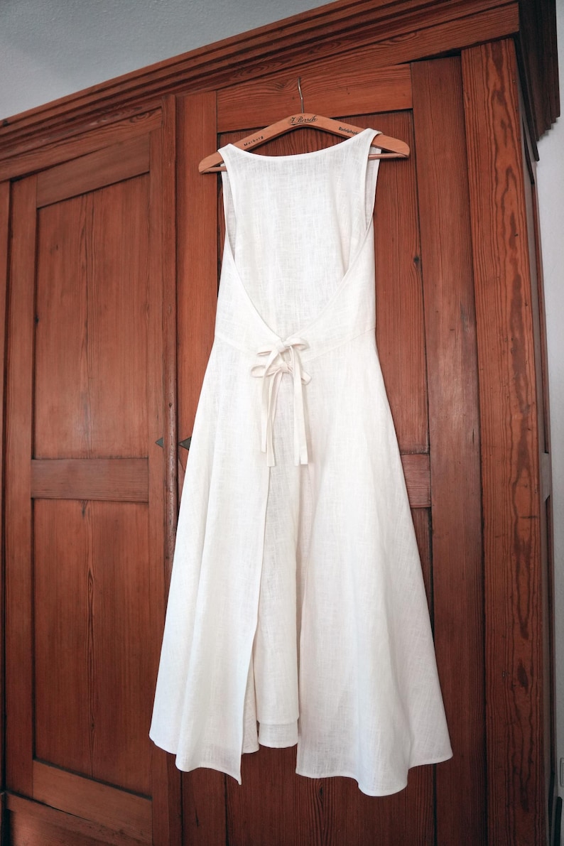 May include: A sleeveless off-white dress featuring a deep open back design with a tie closure at the waist. The dress has a flared skirt that falls below the knee and is constructed from a textured fabric, hanging from a wooden clothes hanger.