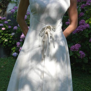 May include: A sleeveless off-white dress featuring a high boat neckline and a fitted bodice. The waist is defined by a horizontal seam and a matching fabric tie closure centered at the front. The skirt flows into a full, floor-length silhouette.
