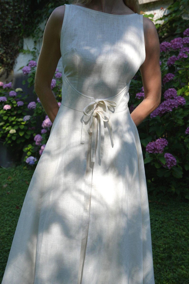 May include: A sleeveless off-white dress featuring a high boat neckline and a fitted bodice. The waist is defined by a horizontal seam and a matching fabric tie closure centered at the front. The skirt flows into a full, floor-length silhouette.