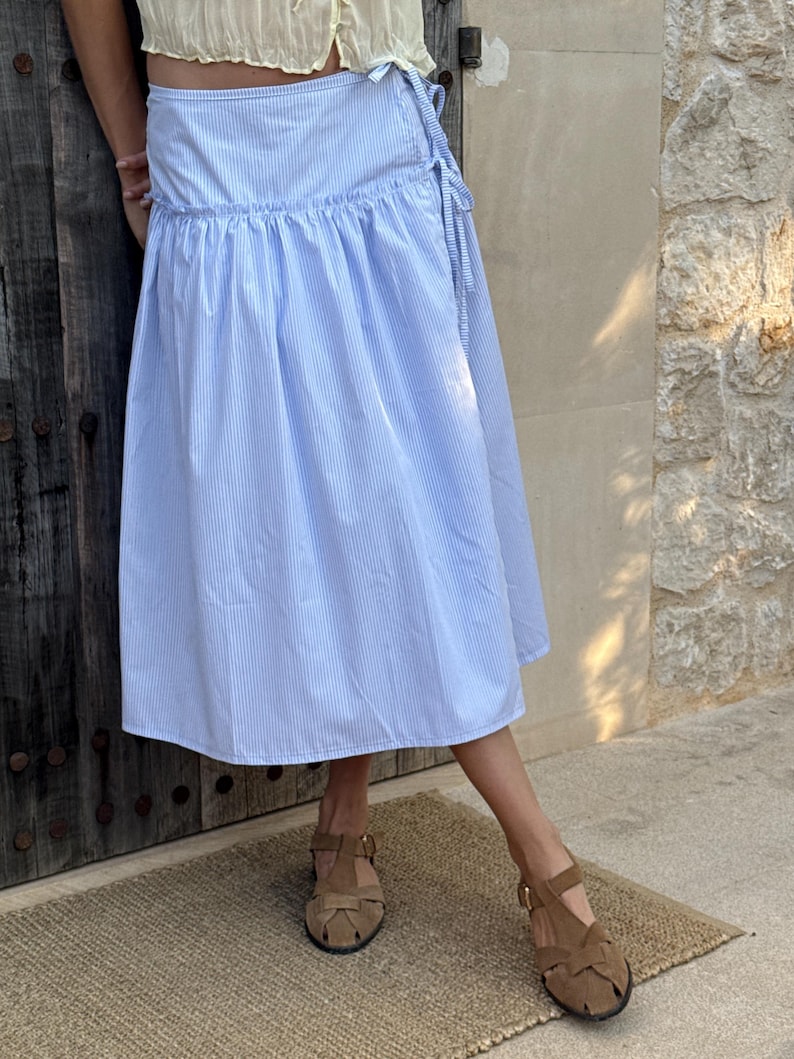 Puede incluir: Una falda envolvente a rayas azules y blancas con cintura fruncida. La falda se combina con sandalias marrones. La falda es de largo midi y parece estar hecha de una tela ligera.