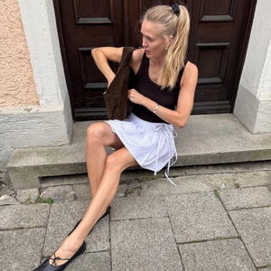 Puede incluir: Una mujer con un top negro, una falda blanca de lunares y bailarinas negras. Sostiene un bolso de ante marr&oacute;n. El fondo muestra una puerta de madera oscura y un escal&oacute;n de piedra.