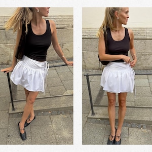 Puede incluir: Una camiseta sin mangas negra combinada con una minifalda blanca. La falda presenta un estampado de rayas y detalles de cord&oacute;n. El atuendo se completa con bailarinas negras y un peque&ntilde;o bolso negro. El fondo es una pared de piedra y una acera.