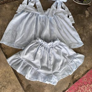 May include: A pale blue two-piece outfit, comprising a top with tie straps and a ruffled hem, and matching ruffled shorts. The fabric has a textured look. The outfit is displayed on a neutral surface.