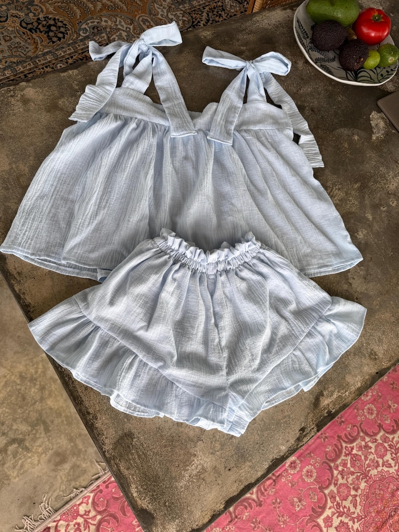 May include: A pale blue two-piece outfit, comprising a top with tie straps and a ruffled hem, and matching ruffled shorts. The fabric has a textured look. The outfit is displayed on a neutral surface.