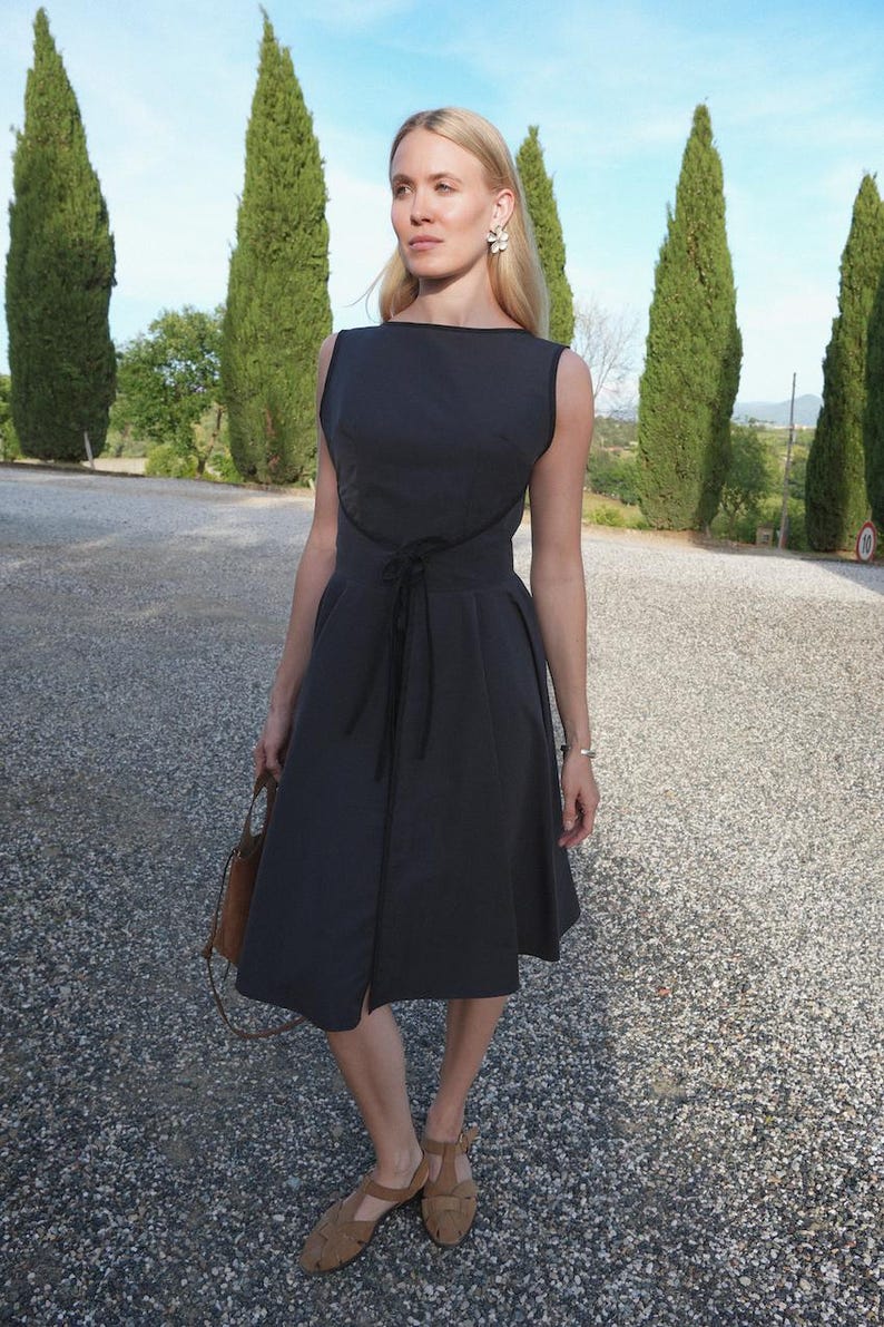 May include: A sleeveless charcoal gray midi dress featuring a boat neckline, a fitted bodice with a defined waistline, and a flared skirt. The dress includes a thin black tie closure at the center waist and black piping trim along the neckline and armholes.