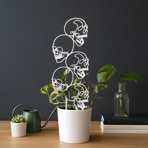 Peut inclure: Un tuteur de plante blanc avec cinq silhouettes de crânes empilés. Il est placé dans un pot blanc avec des plantes vertes. Un petit succulent et un arrosoir vert sont à gauche.