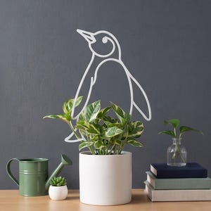 以下が含まれることがあります： ペンギンのシルエットをかたどった白いワイヤー製のプラントトレリス。緑と白の斑入りのポトスが植えられた白い円筒形のセラミック製植木鉢に差し込まれており、他の装飾品と共に木製の表面の上に置かれています。