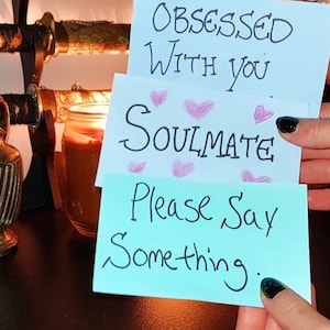 Op de afbeelding: Drie lichtblauwe kaarten met handgeschreven tekst: "Obsessed With You", "Soulmate" met roze hartjes en "Please Say Something". De kaarten worden voor een kaars en decoratieve objecten gehouden.