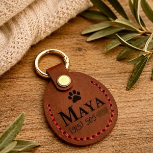 May include: A round brown leather pet tag featuring a gold-tone metal keyring, a brass rivet, and red stitching along the edge. The tag is engraved with a black paw print icon, the name MAYA, and the phone number (915) 505- followed by blurred digits.