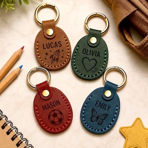 May include: Four oval-shaped leather keychains with gold-tone metal rings and rivets. Each keychain features a unique color: tan, green, red, and blue. They are engraved with names and icons: Lucas with a rocket, Olivia with a heart, Mason with a soccer ball, and Emily with a butterfly. Each piece has visible perimeter stitching.