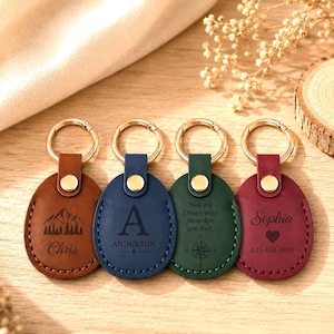 May include: Four oval-shaped leather keychains in brown, blue, green, and maroon, each with a gold-tone metal ring and snap button. The brown keychain features a mountain graphic and the name Chris. The blue one displays the letter A, the name Anderson, and a star. The green one shows a compass and the text Not all those who wander are lost. The maroon one displays the name Sophia, a heart, and the number 123-456-7890.