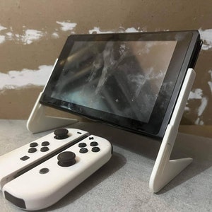 May include: A white Nintendo Switch console with a white stand and two white Joy-Con controllers. The console is in tablet mode and the screen is displaying a blurry image.