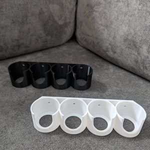 Könnte beinhalten: Zwei 3D-gedruckte Wandhalterungen zum Aufbewahren von Gegenständen, eine schwarze und eine weiße. Jede Halterung hat vier runde Öffnungen.