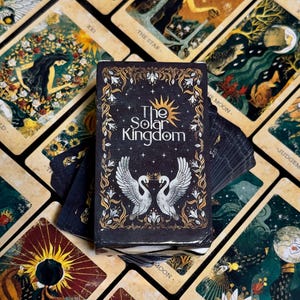 Könnte beinhalten: Ein Tarotkartenspiel mit einer schwarzen Schachtel mit dem Text "The Solar Kingdom" und zwei weißen Schwänen. Die Karten zeigen Illustrationen mit himmlischen und natürlichen Themen, darunter Mond, Sonne und Sterne. Die Karten sind im Vintage-Stil gehalten.