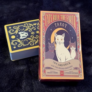 Può includere: Un mazzo di carte dei tarocchi intitolato "Cats Rule the Earth" con una scatola che presenta illustrazioni di tre gatti. Il mazzo include 78 carte ed è pensato per gli appassionati di felini. La scatola è arancione, rossa e color crema.