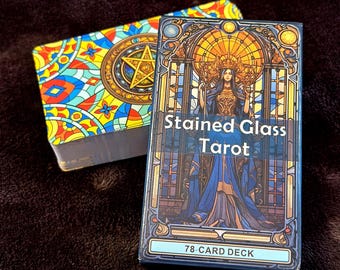 May include: A Stained Glass Tarot 78-card deck box featuring a figure in a blue gown against a stained glass window design, with the text Stained Glass Tarot. Beside it lies a second deck with a colourful geometric pattern and a gold pentacle.