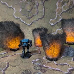 May include: A miniature black and blue mech robot stands on a hexagonal game board, surrounded by four dark, fluffy, fiery-looking terrain pieces. The board has a map design with brown and green tones.