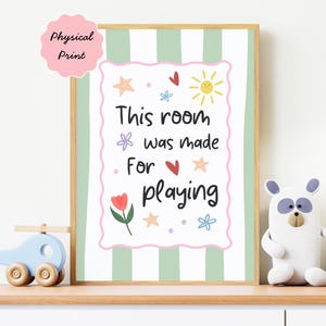 May include: A framed print with the text "This room was made for playing" in black script, surrounded by colorful illustrations. The background has green and white stripes. A pink sticker in the upper left corner says "Physical Print".