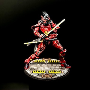 May include: A red and black painted miniature figure of a samurai warrior holding a katana sword. The figure is standing on a circular base with a textured surface.