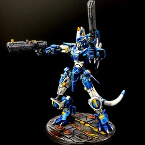 May include: A blue and white robot with a yellow and black gun, standing on a circular base with a black and yellow design.