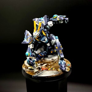 May include: A blue and yellow painted robot figurine with a detailed design. The robot is posed in a dynamic action pose with one arm raised and the other arm bent at the elbow. The robot is standing on a base that is painted to look like a destroyed city street.