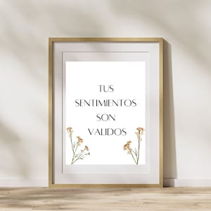 May include: A framed print with a white background and black text that reads "Tus Sentimientos Son Validos". The text is centered on the page and there are small dried flowers in the corners of the print.