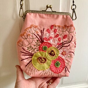 May include: A pink fabric purse with a floral embroidery design. The purse has a clasp closure and a chain strap.