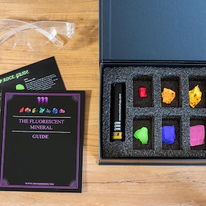 Op de afbeelding: Een zwarte doos met een set van zes fluorescerende mineralen, een UV-lamp en een handleiding. De handleiding heeft de titel "The Fluorescent Mineral Guide" en de website "www.miner shop.com".