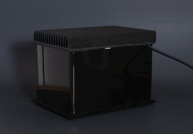 Puede incluir: Un dispositivo electr&oacute;nico negro y rectangular con una parte superior acanalada y una superficie brillante y reflectante. Un cable negro se extiende desde el lateral. El dispositivo est&aacute; sobre un fondo gris oscuro.