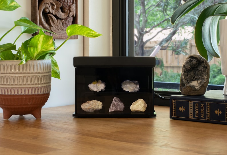 Puede incluir: Una vitrina negra con una selecci&oacute;n de cristales y minerales. La vitrina est&aacute; sobre una superficie de madera, con una planta en maceta y una piedra decorativa cerca. Los cristales son de varios colores y formas.