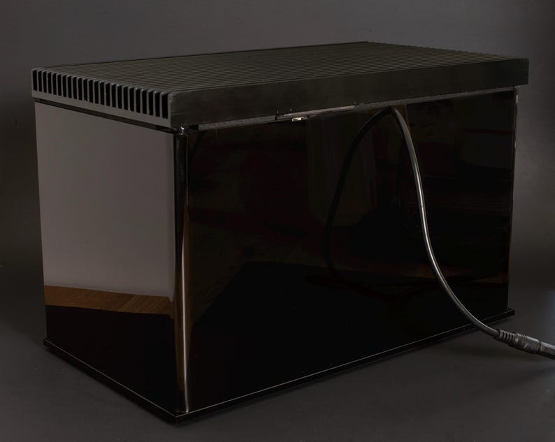 May include: A black rectangular box with a black cord attached to it. The box has a vent on the top and a glossy finish.