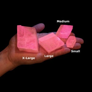 May include: Four translucent, bright pink crystalline mineral specimens of varying sizes labeled X-Large, Large, Medium, and Small. The geometric, angular stones have a vibrant, glowing appearance against a solid black background, showcasing distinct facets.