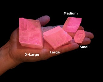 May include: Four translucent, bright pink crystalline mineral specimens of varying sizes labeled X-Large, Large, Medium, and Small. The geometric, angular stones have a vibrant, glowing appearance against a solid black background, showcasing distinct facets.