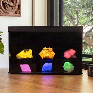 May include: A black display case with a glass front showcasing six different colored rocks illuminated with colored lights. The rocks are yellow, pink, green, blue, and purple. The case is sitting on a wooden table.