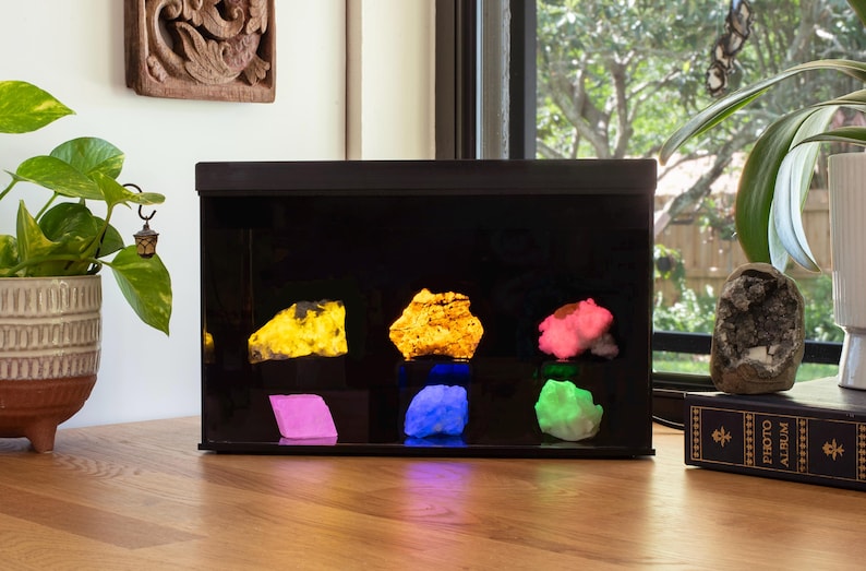 May include: A black display case with a glass front showcasing six different colored rocks illuminated with colored lights. The rocks are yellow, pink, green, blue, and purple. The case is sitting on a wooden table.