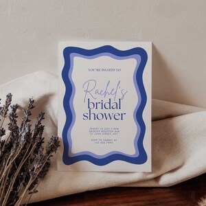 Op de afbeelding: Een blauwe en witte uitnodigingskaart met een golvende rand. De tekst op de kaart luidt "You're invited to Rachel's bridal shower. Friday 12 July 7-9pm Groove Rooftop Bar 31 Love Street, NYI RSVP to Sammy at 123 456.7890"
